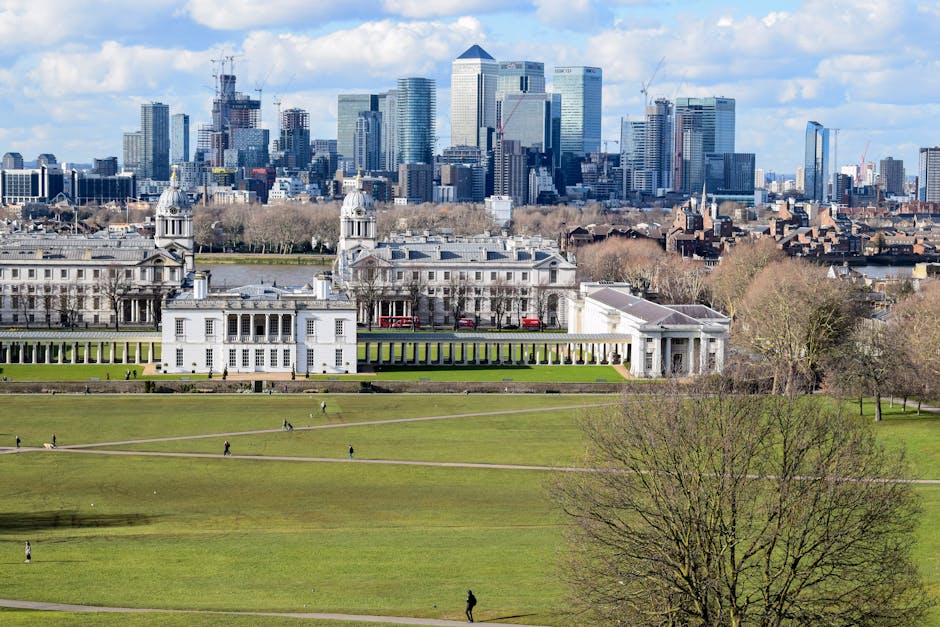 A scene of two individuals sitting on a park bench beneath a large leafy tree near the river, with a view of historic Greenwich buildings and a prominent dome in the background. The bench is positione