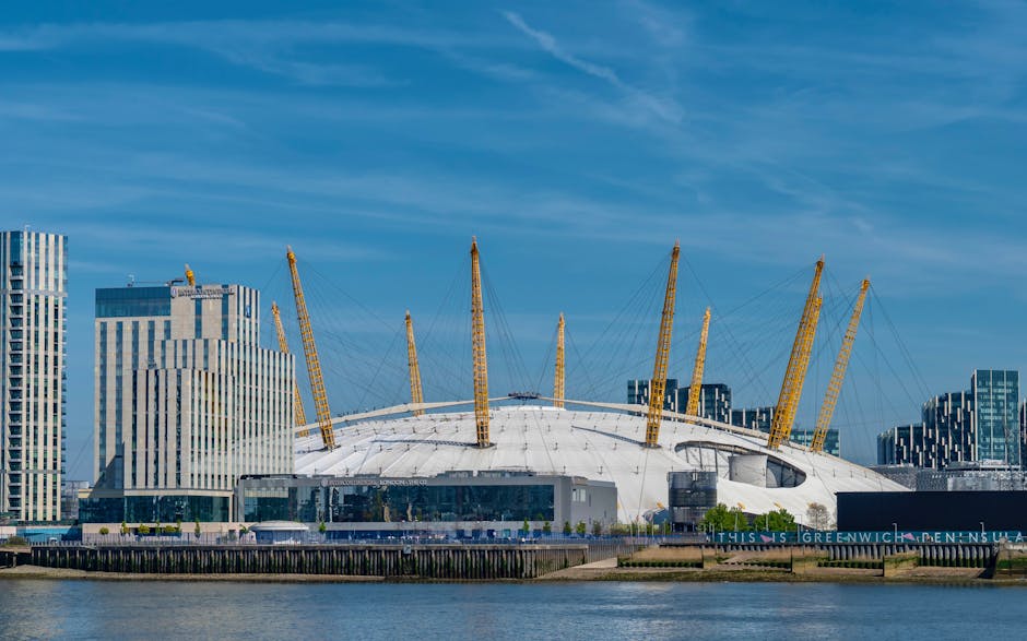 A large historic building with a domed clock tower in Greenwich, London, featuring white stone architecture with classical columns and ornate details, set against a clear blue sky. In the foreground, 