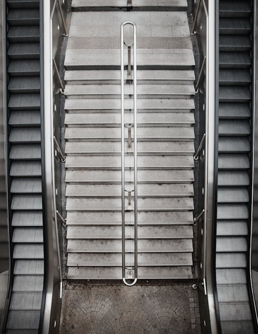 Inside a modern indoor stairwell with metal railings and yellow safety strips on each step, leading up to a bright opening with natural light filtering through. The staircase has a central handrail an