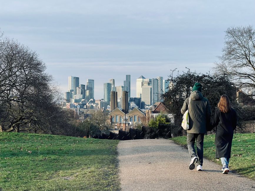 A wide urban skyline of modern high-rise buildings and skyscrapers in Greenwich, London, with some under construction cranes visible atop the tallest structures. In the foreground, there is greenery i